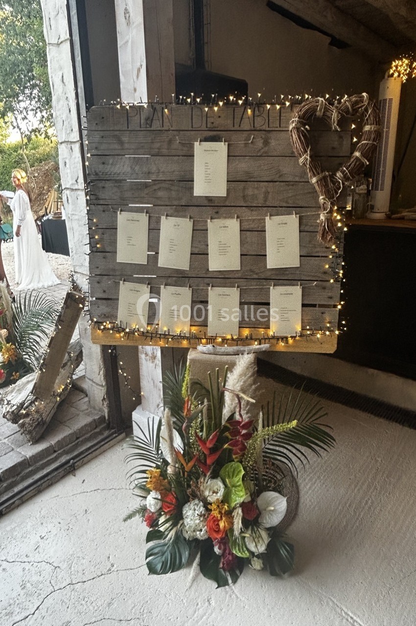 Plan de table affiché sur une palette en bois décorée de guirlandes lumineuses, avec un bouquet floral en bas.