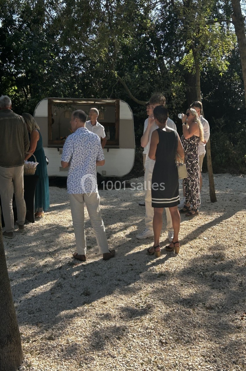 Groupe de personnes discutant devant une caravane aménagée en stand, dans un espace extérieur ombragé.