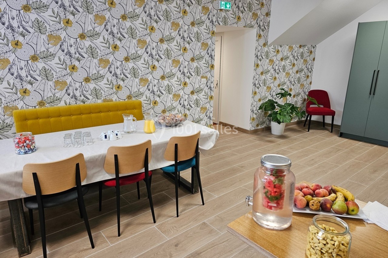 Salle lumineuse avec table dressée, chaises colorées, carafe d'eau infusée, fruits et décor floral sur les murs.