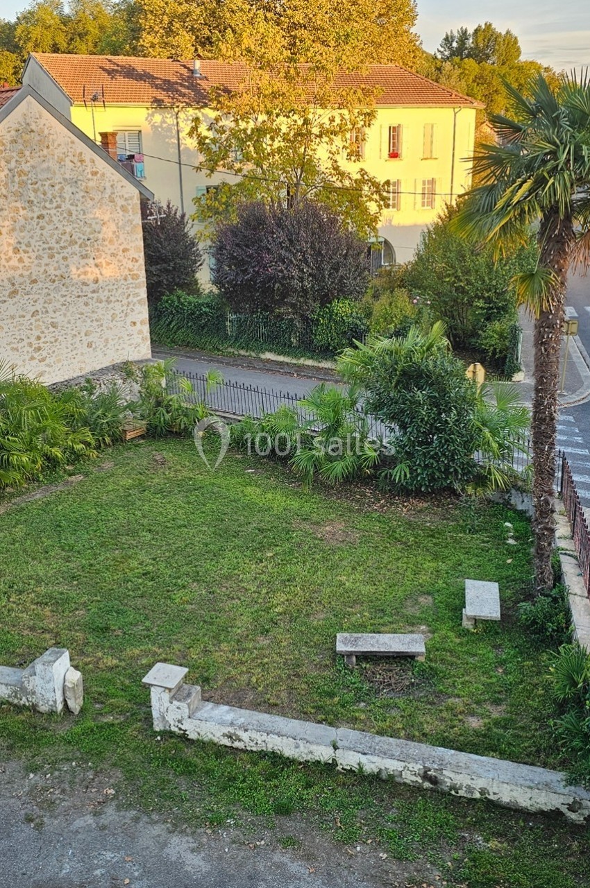 Jardin avec pelouse, bancs en pierre, palmiers et arbres, entouré de bâtiments en pierre et végétation.