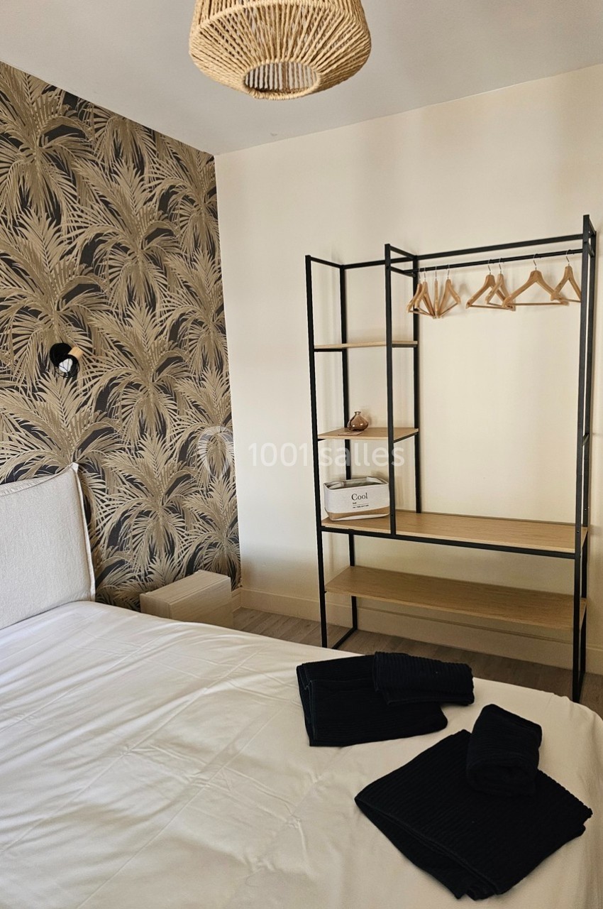 Chambre avec lit blanc, portant en métal noir avec cintres, mur à motif végétal et suspension en rotin.