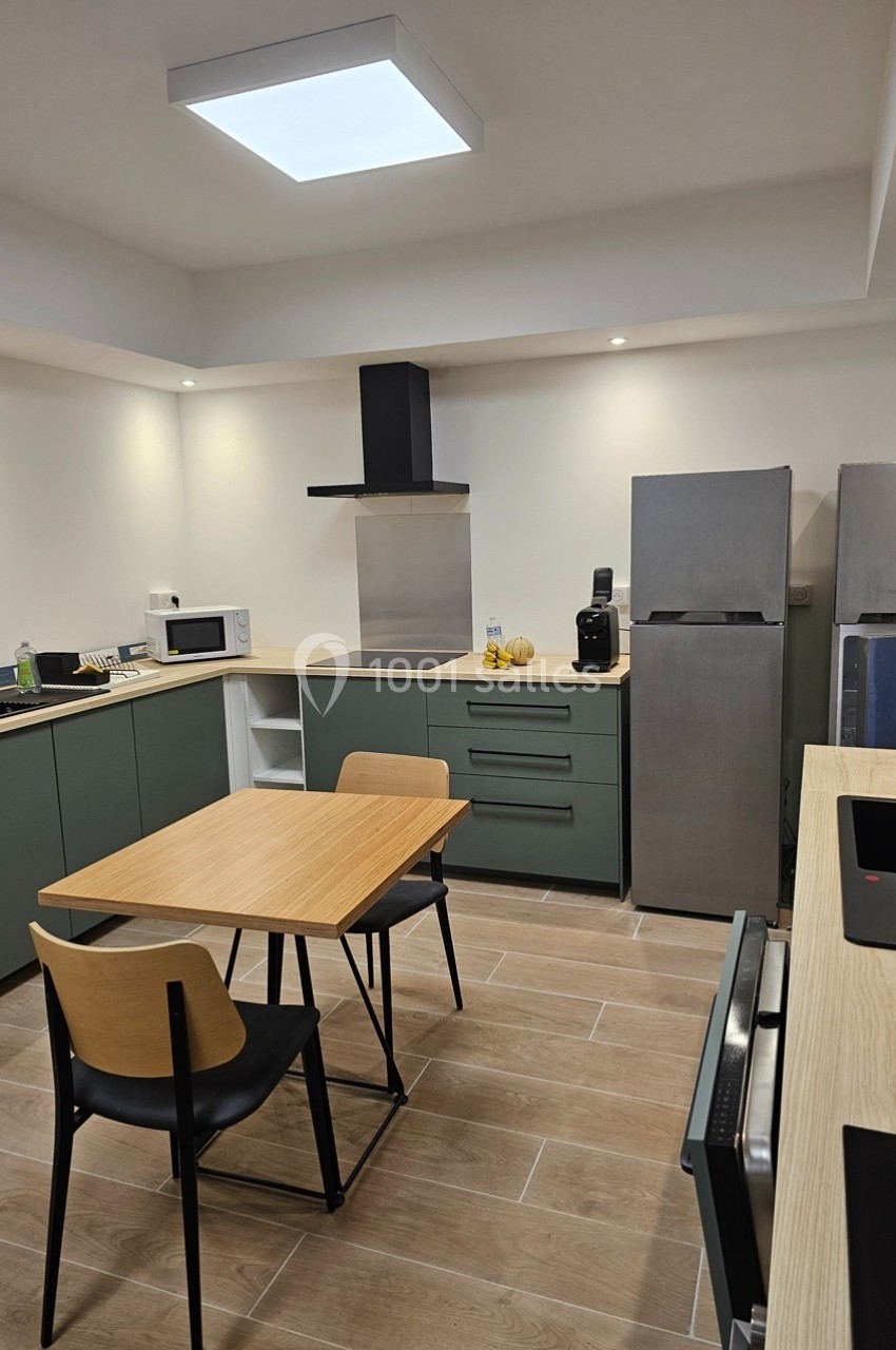 Cuisine moderne avec meubles verts, table en bois, chaises noires, électroménagers en acier et éclairage au plafond.