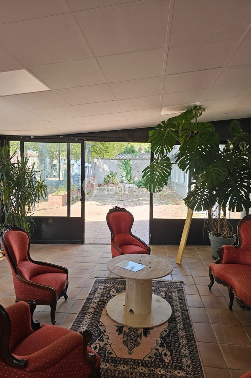Salon lumineux avec fauteuils rouges, table basse en bois, plantes vertes et vue sur un jardin extérieur.