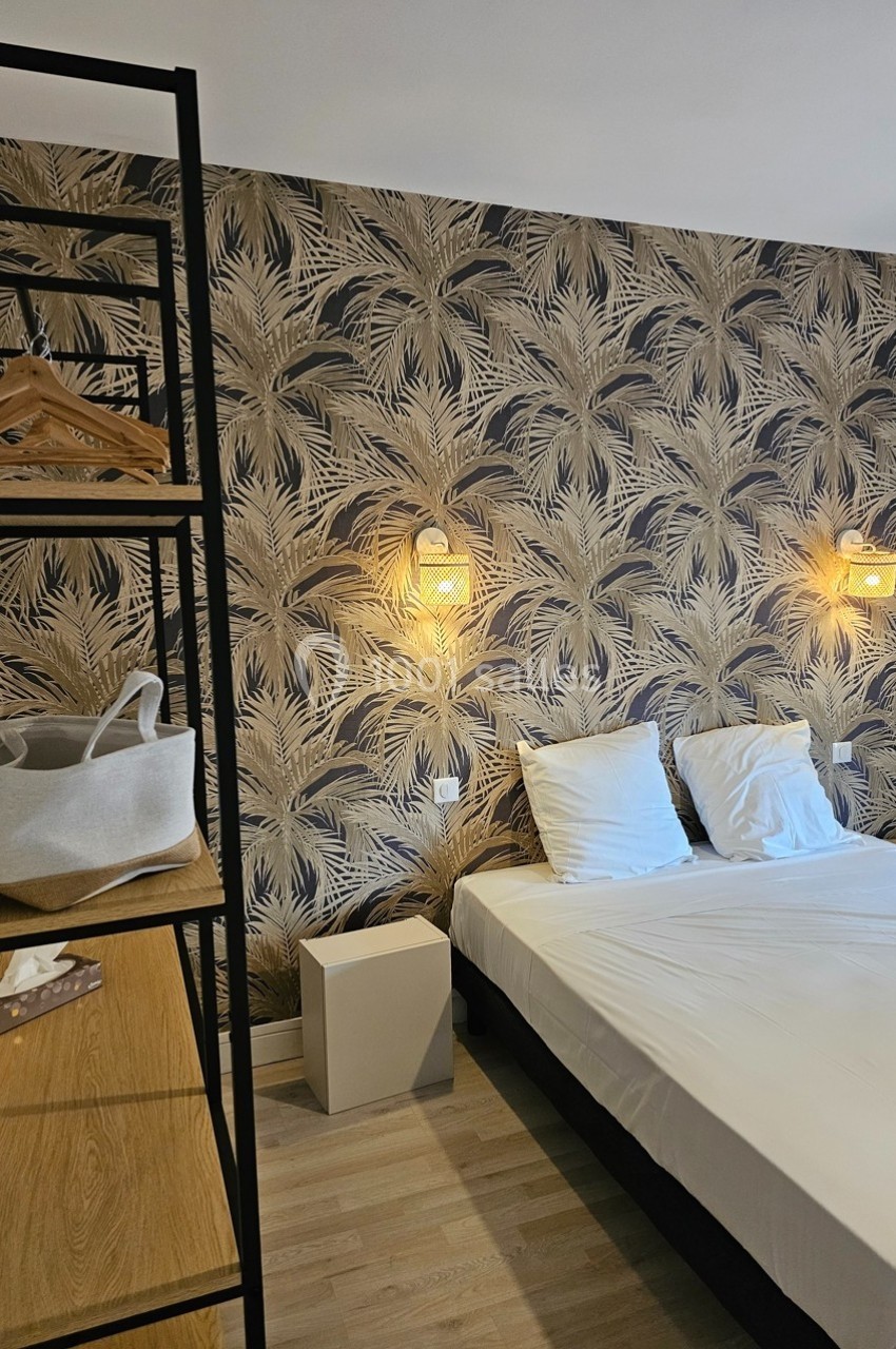 Lit double avec draps blancs, éclairé par deux appliques murales, devant un mur à motif végétal, étagère à gauche.