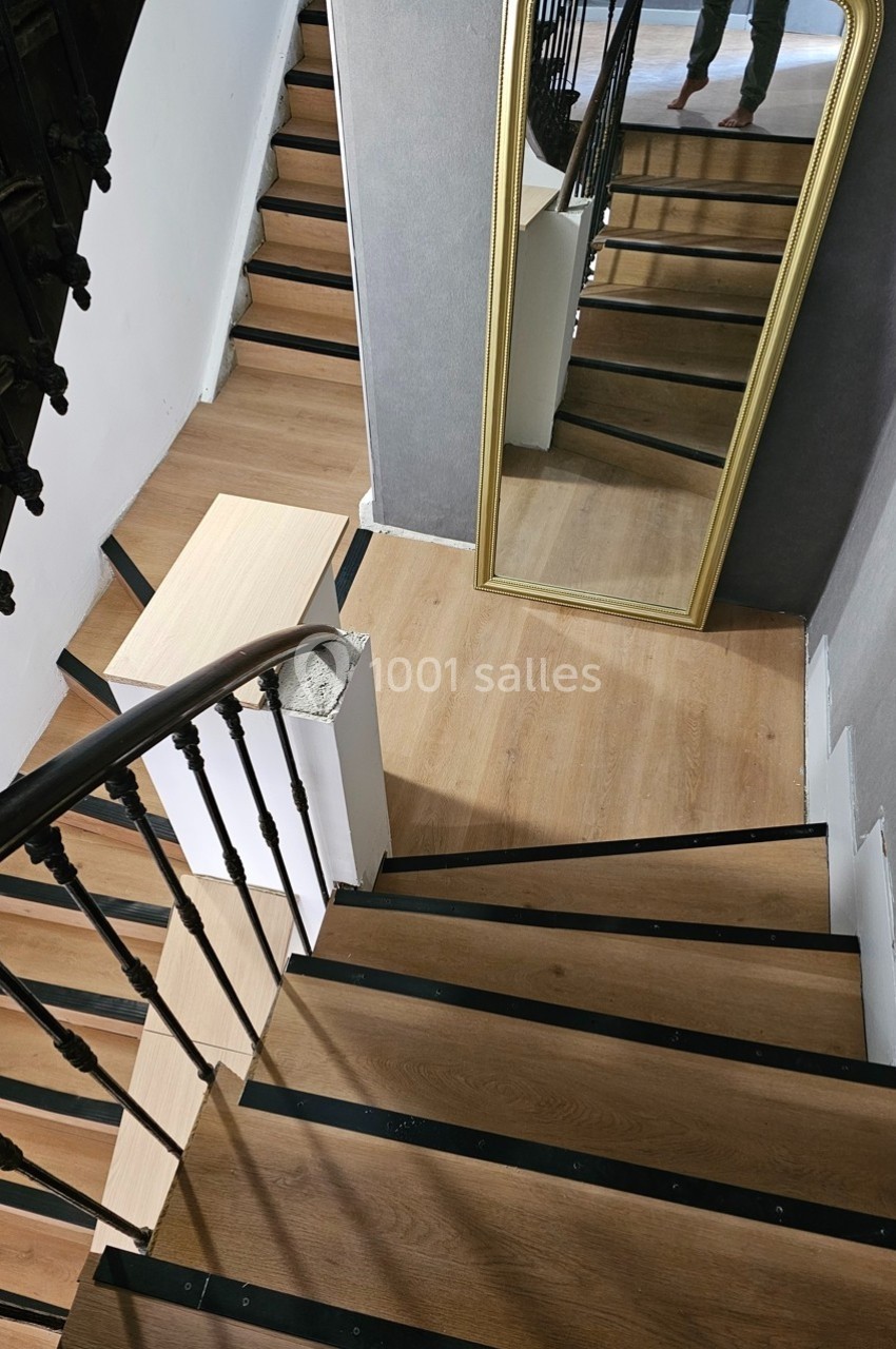 Escalier en bois avec rampe en métal noir, grand miroir doré appuyé contre un mur gris.