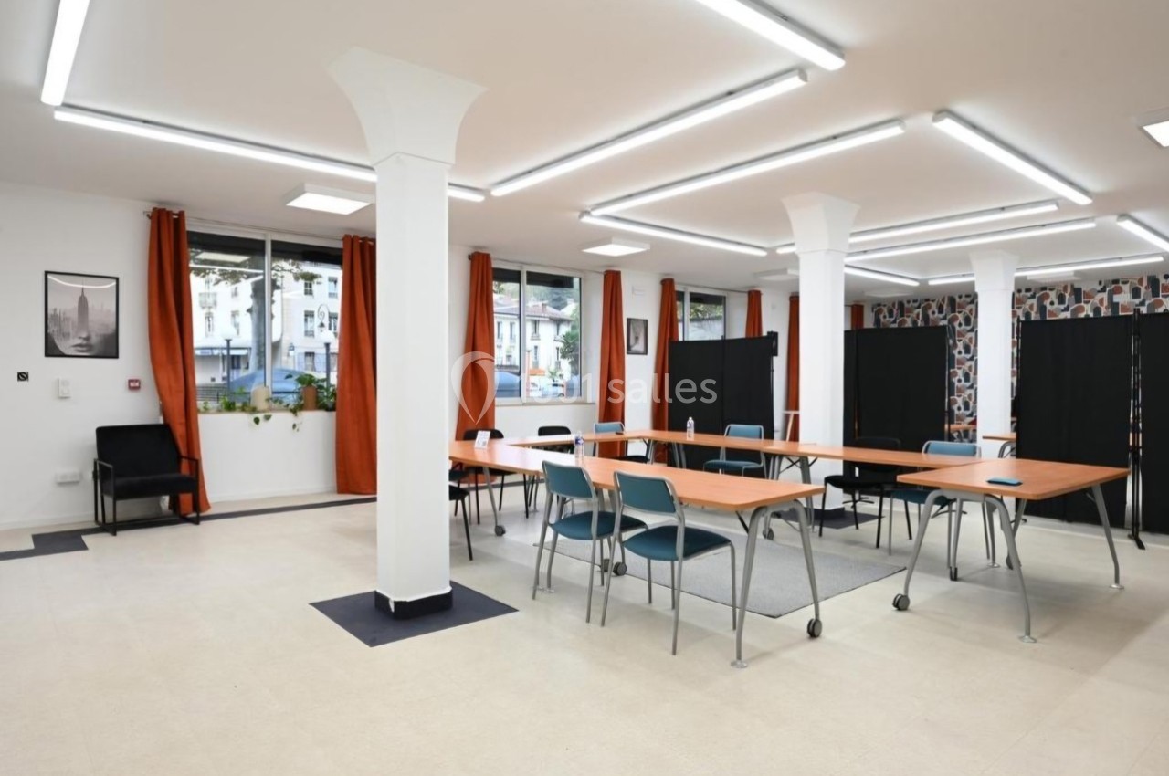 Salle lumineuse avec grandes fenêtres, tables en bois disposées en U, chaises et rideaux orange.