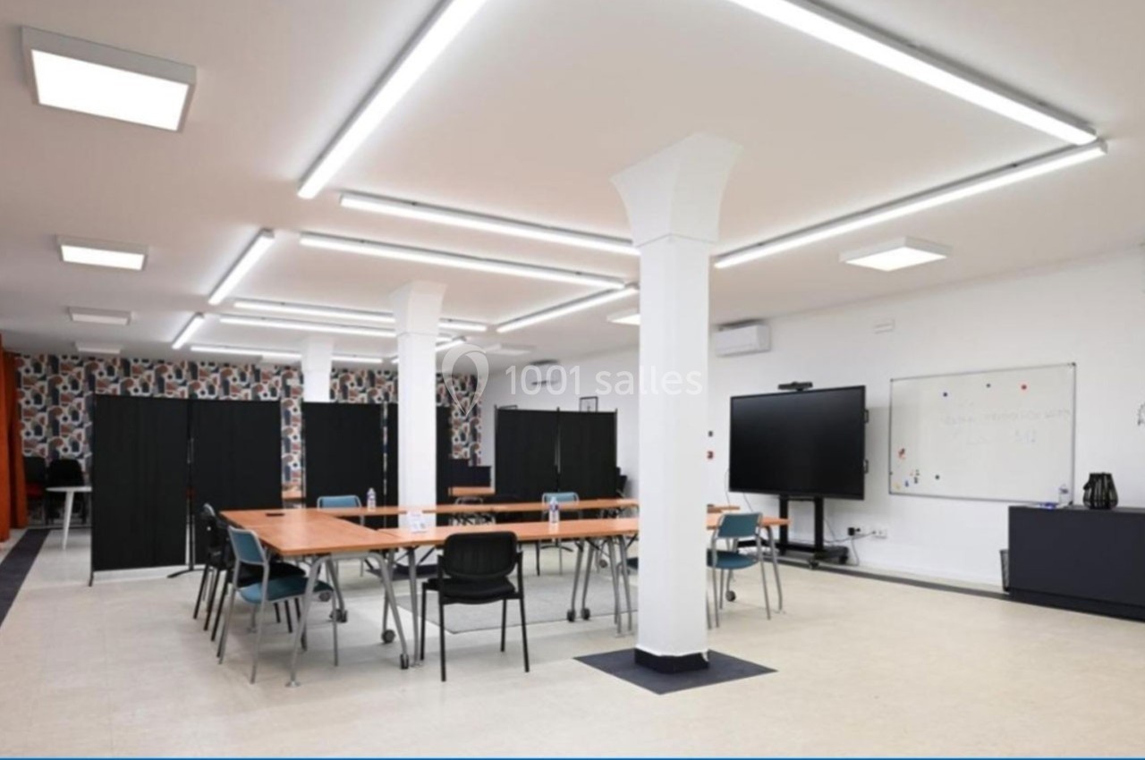 Salle lumineuse avec tables en bois, chaises, tableau blanc, écran et panneaux de séparation noirs.