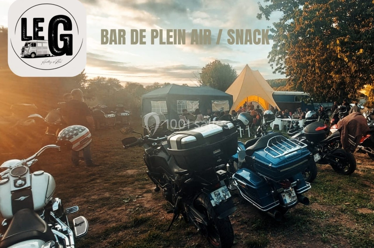 Rassemblement de motos dans un espace extérieur avec tentes, arbres et lumière de fin de journée.