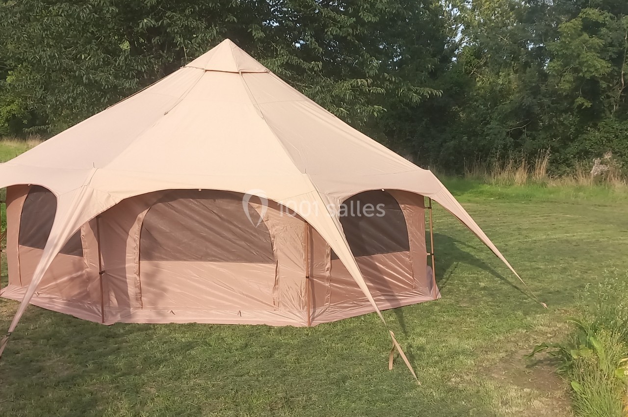 Tente beige de grande taille montée sur une pelouse, entourée d'arbres et de végétation.