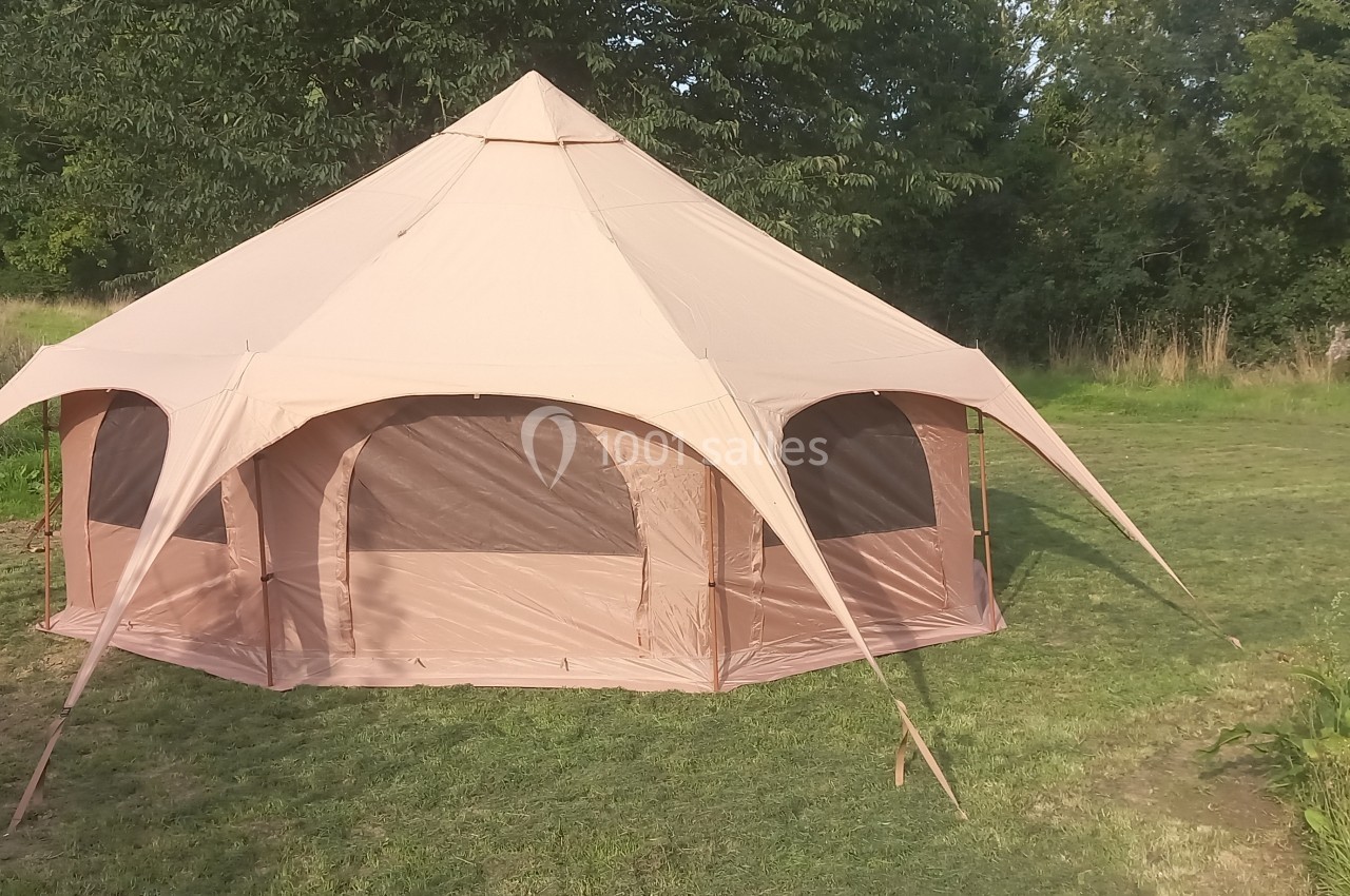 Tente beige en forme de tipi montée sur une pelouse, entourée de végétation et d'arbres.