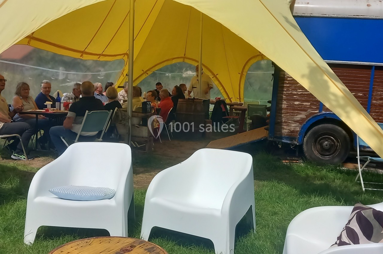 Groupe de personnes assises sous une tente jaune, près d'un bus bleu, entourées de chaises et de tables sur une pelouse.