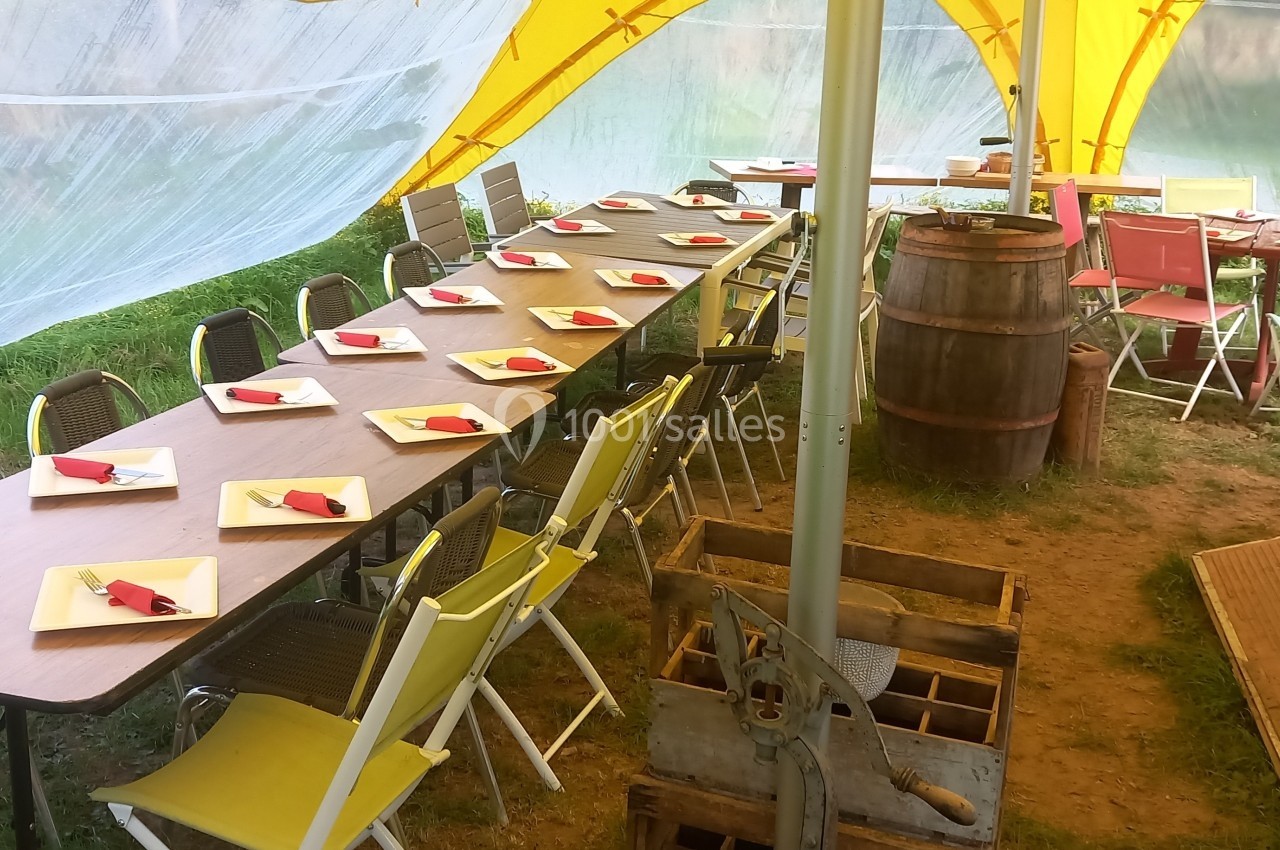Tables dressées avec des assiettes et serviettes rouges sous une tente jaune, entourées de chaises dans un cadre extérieur.