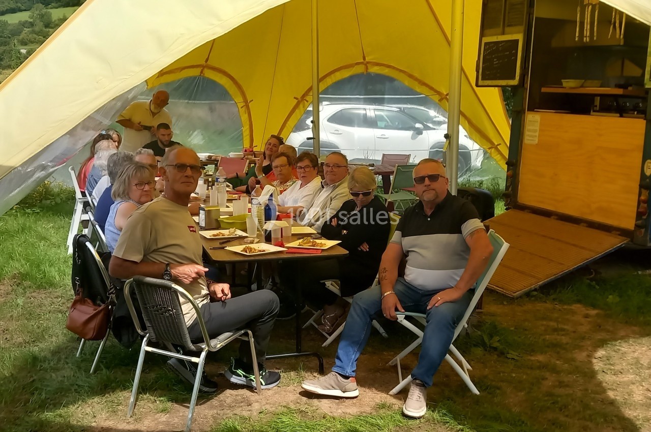 Groupe de personnes assises autour d'une table sous une tente, partageant un repas près d'une remorque aménagée.