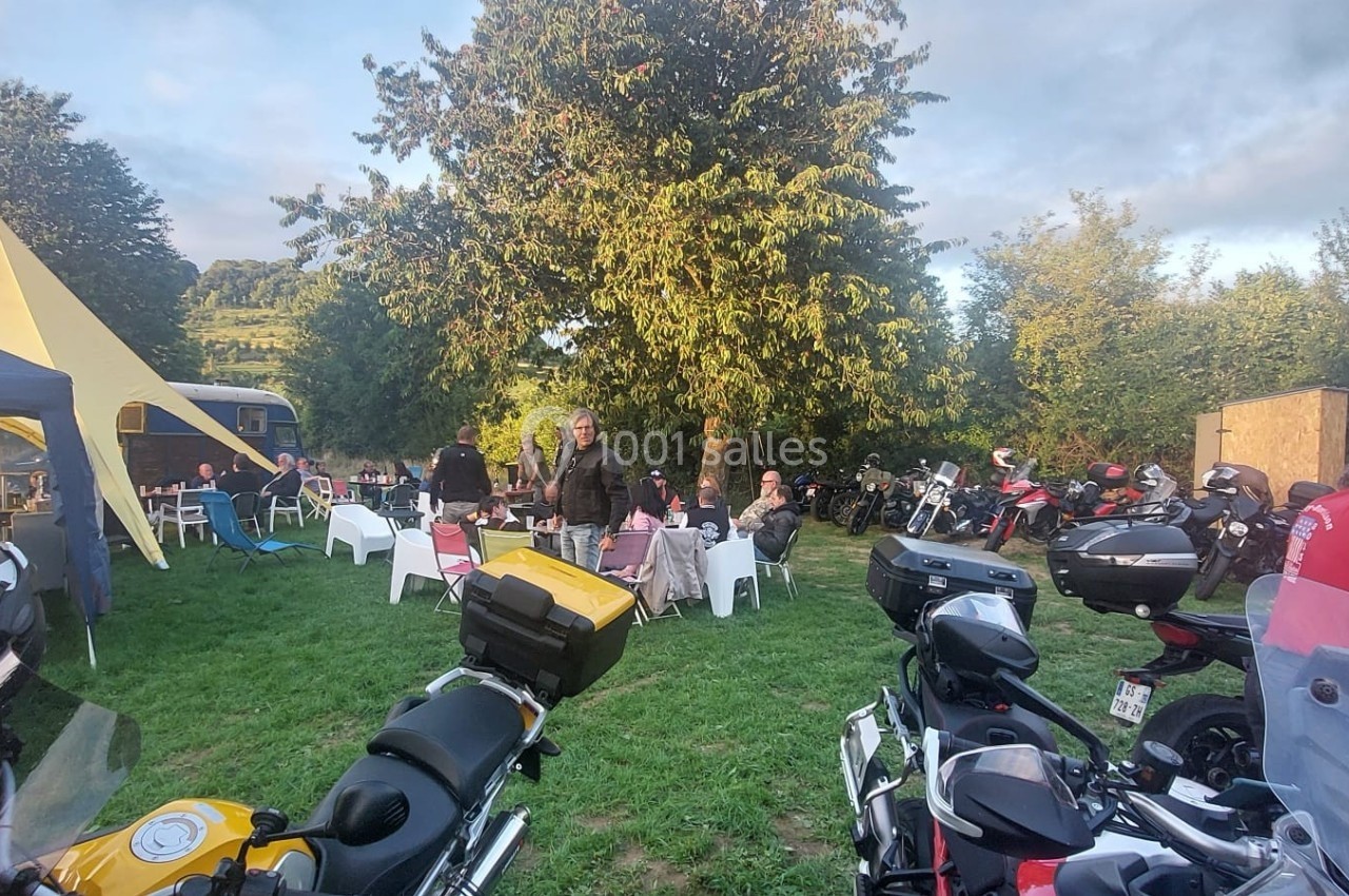 Des motos garées sur une pelouse près de tentes, avec des personnes assises ou debout dans un cadre extérieur arboré.