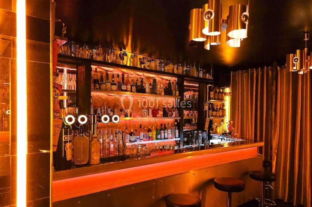 Bar lumineux avec étagères remplies de bouteilles, éclairage orange et doré, tabourets et rideaux en arrière-plan.