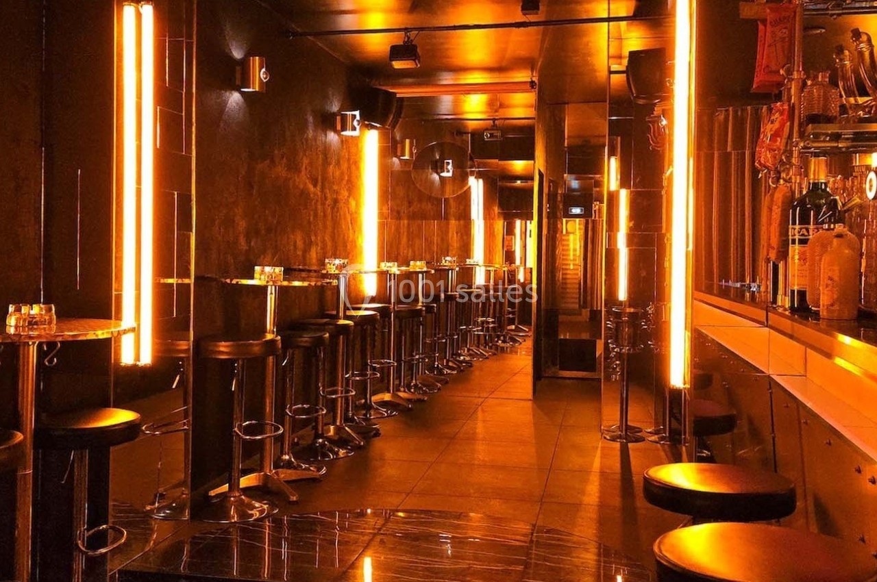 Salle de bar moderne avec éclairage orange néon, tabourets hauts alignés et comptoir à droite.