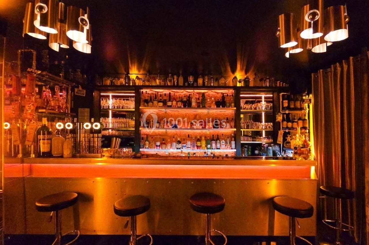 Bar avec comptoir éclairé, tabourets alignés et étagères remplies de bouteilles dans une ambiance tamisée.