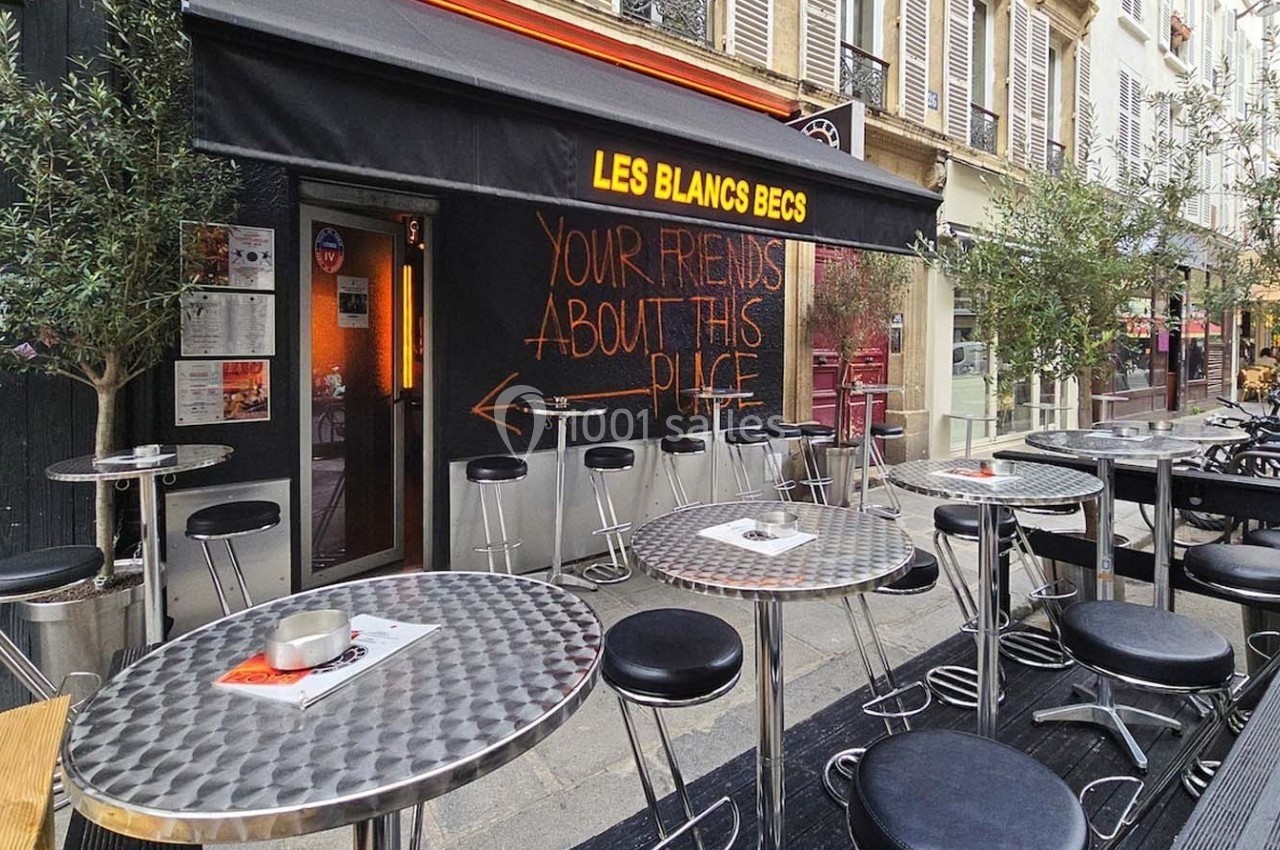 Terrasse d’un café avec tables et chaises métalliques, devanture noire et message écrit sur le mur.