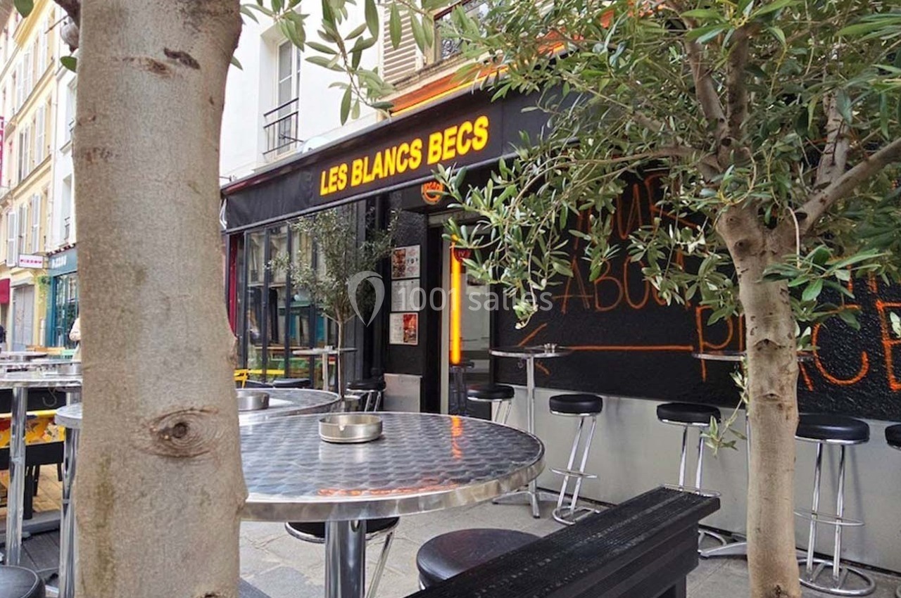 Terrasse extérieure d'un café avec tables en métal, tabourets hauts et enseigne lumineuse ’Les Blancs Becs’.