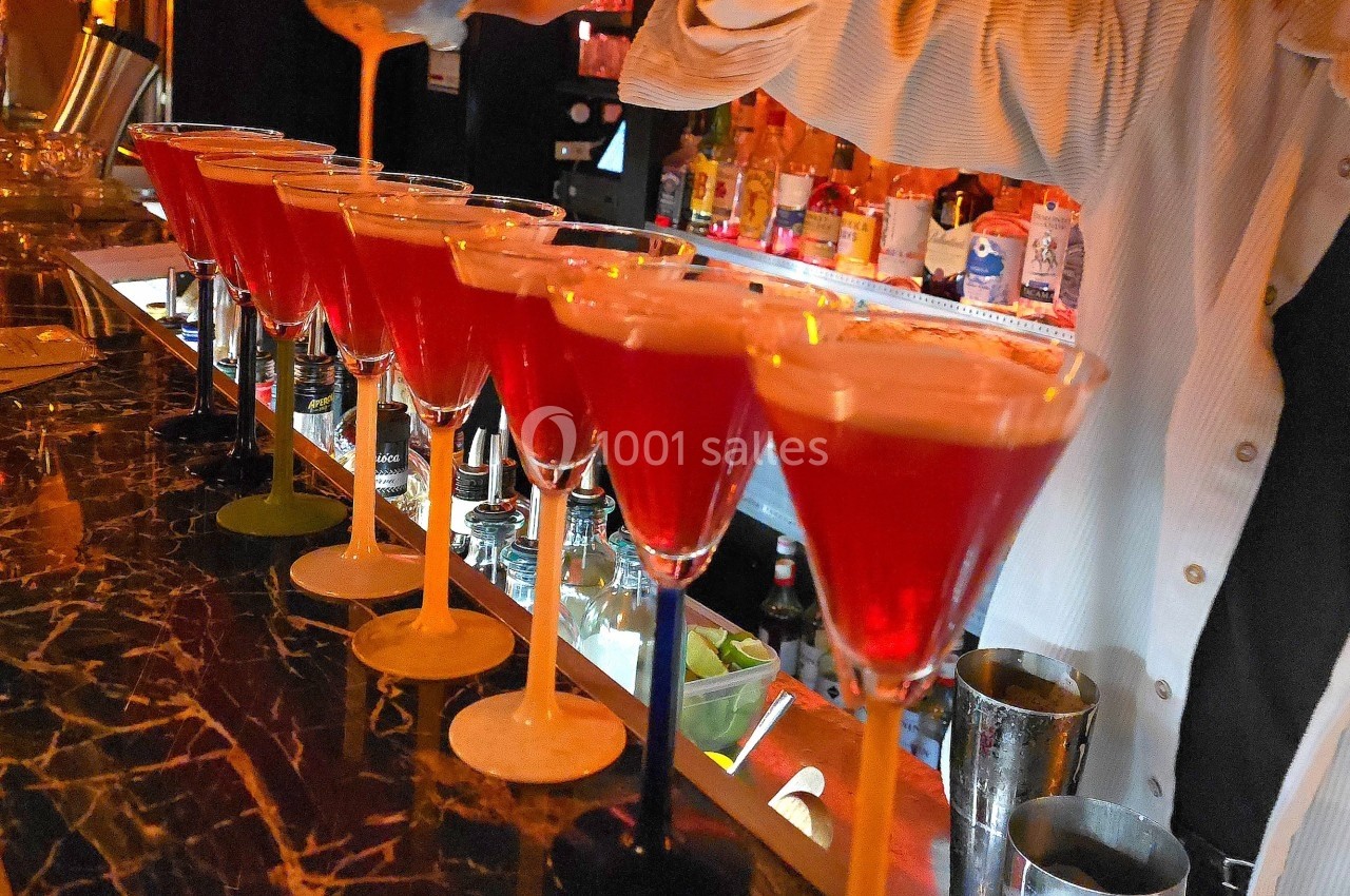 Un barman verse un cocktail rouge dans une rangée de verres alignés sur un comptoir en marbre sombre.