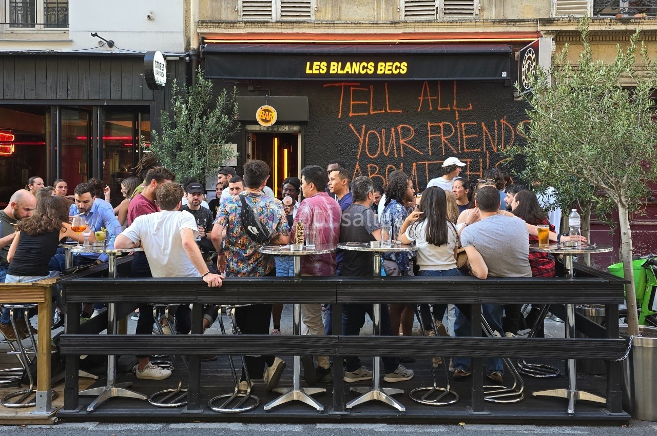 Terrasse animée d'un café avec des clients debout autour de tables hautes, devant une façade noire avec des inscriptions.
