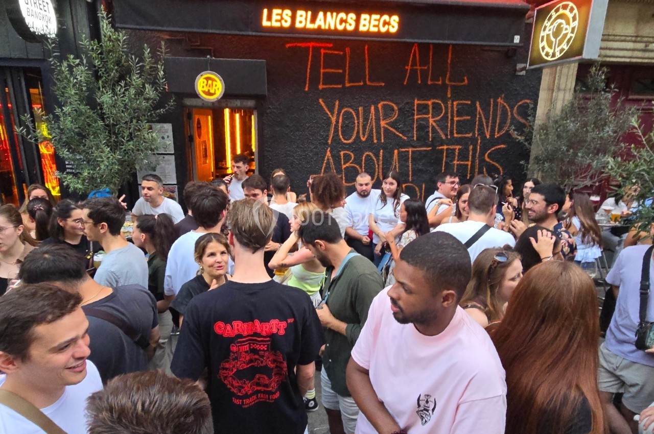 Groupe de personnes rassemblées devant un bar animé, avec une enseigne lumineuse et un graffiti sur le mur.