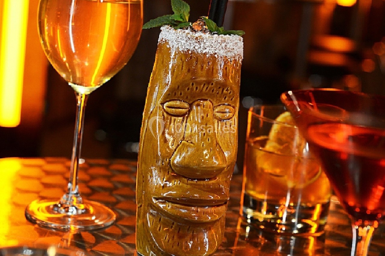 Verre sculpté en forme de visage rempli d'un cocktail, entouré de deux verres de boissons sur une table dorée.