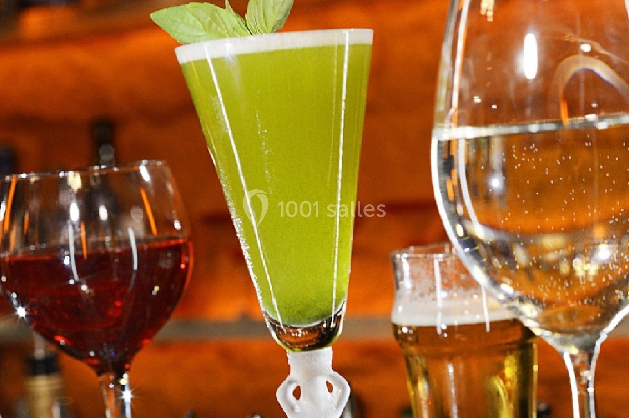 Verres de boissons variées, dont un cocktail vert garni de feuilles, posés sur un bar en arrière-plan flou.