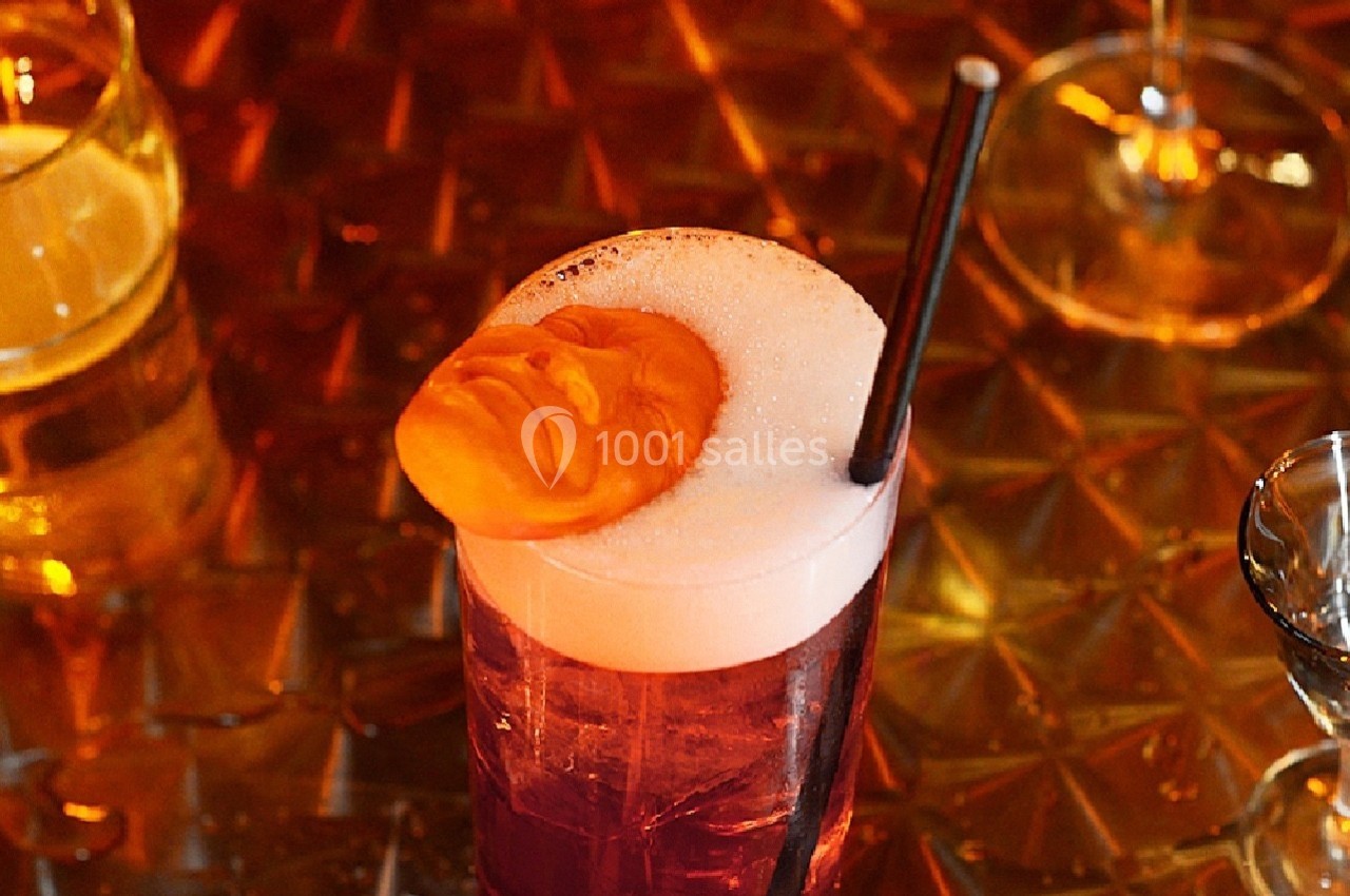 Verre de cocktail rouge avec mousse blanche, décoré d'une figure sculptée orange et une paille noire.
