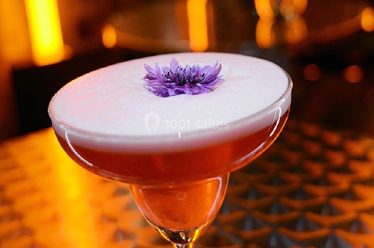 Cocktail rose surmonté d'une mousse blanche et décoré d'une fleur violette, posé sur une table métallique.