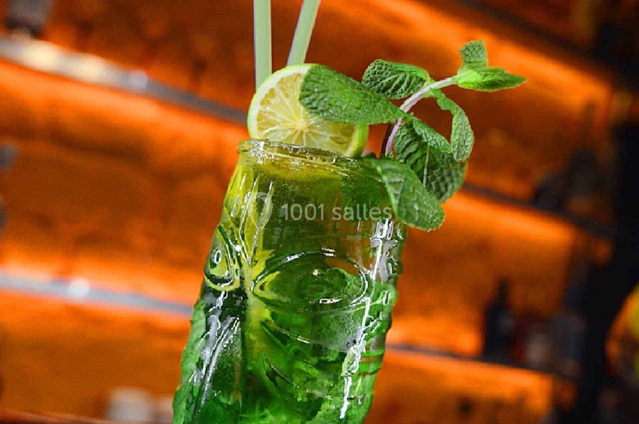 Verre de cocktail vert décoré de menthe fraîche, d'une tranche de citron et de deux pailles, posé sur un bar.