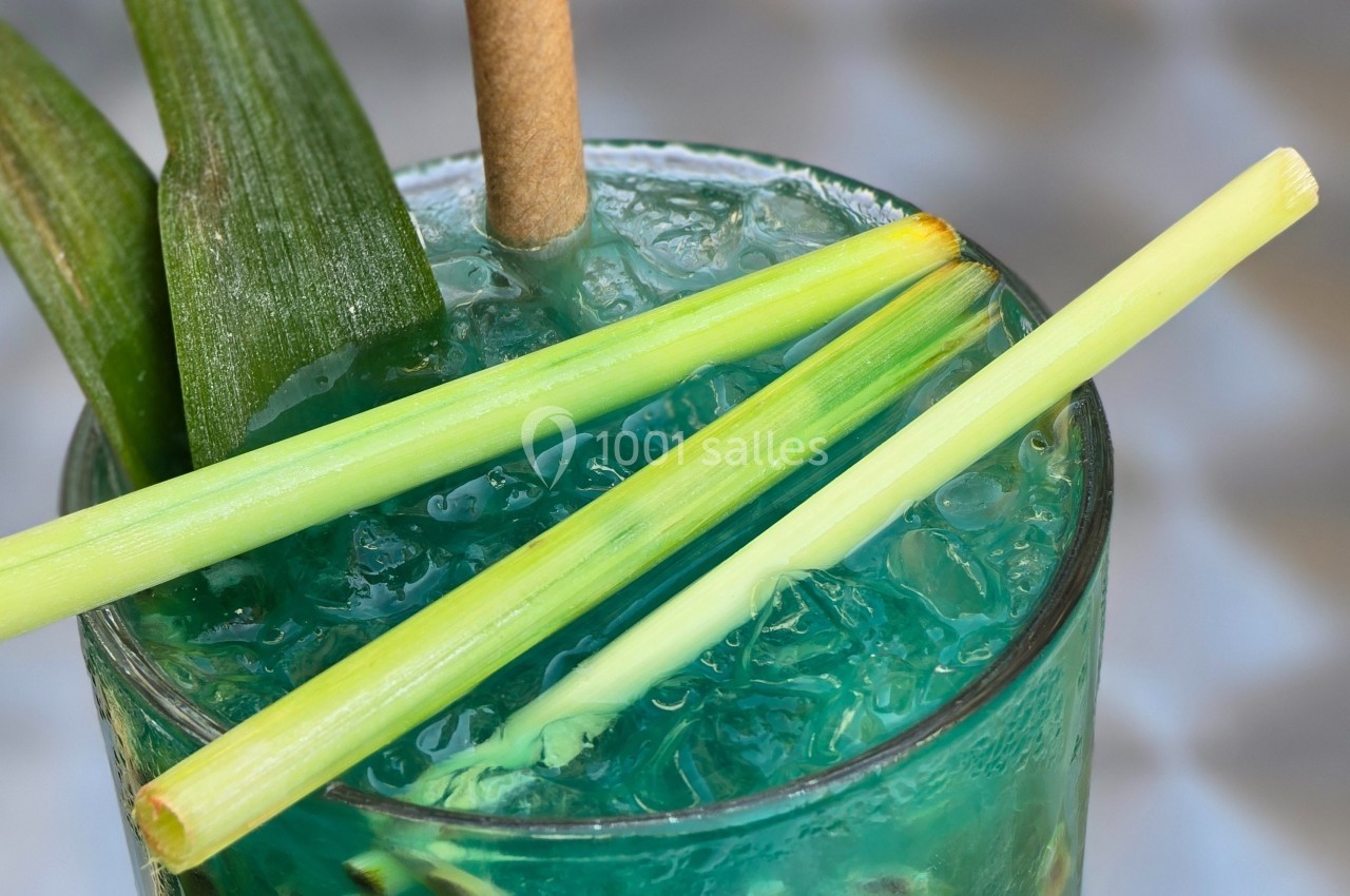 Verre de cocktail bleu avec glaçons, décoré de feuilles vertes et de tiges de citronnelle.