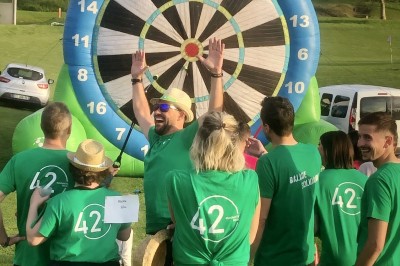 Un groupe de personnes en t-shirts verts participe à une activité de fléchettes géantes en plein air.