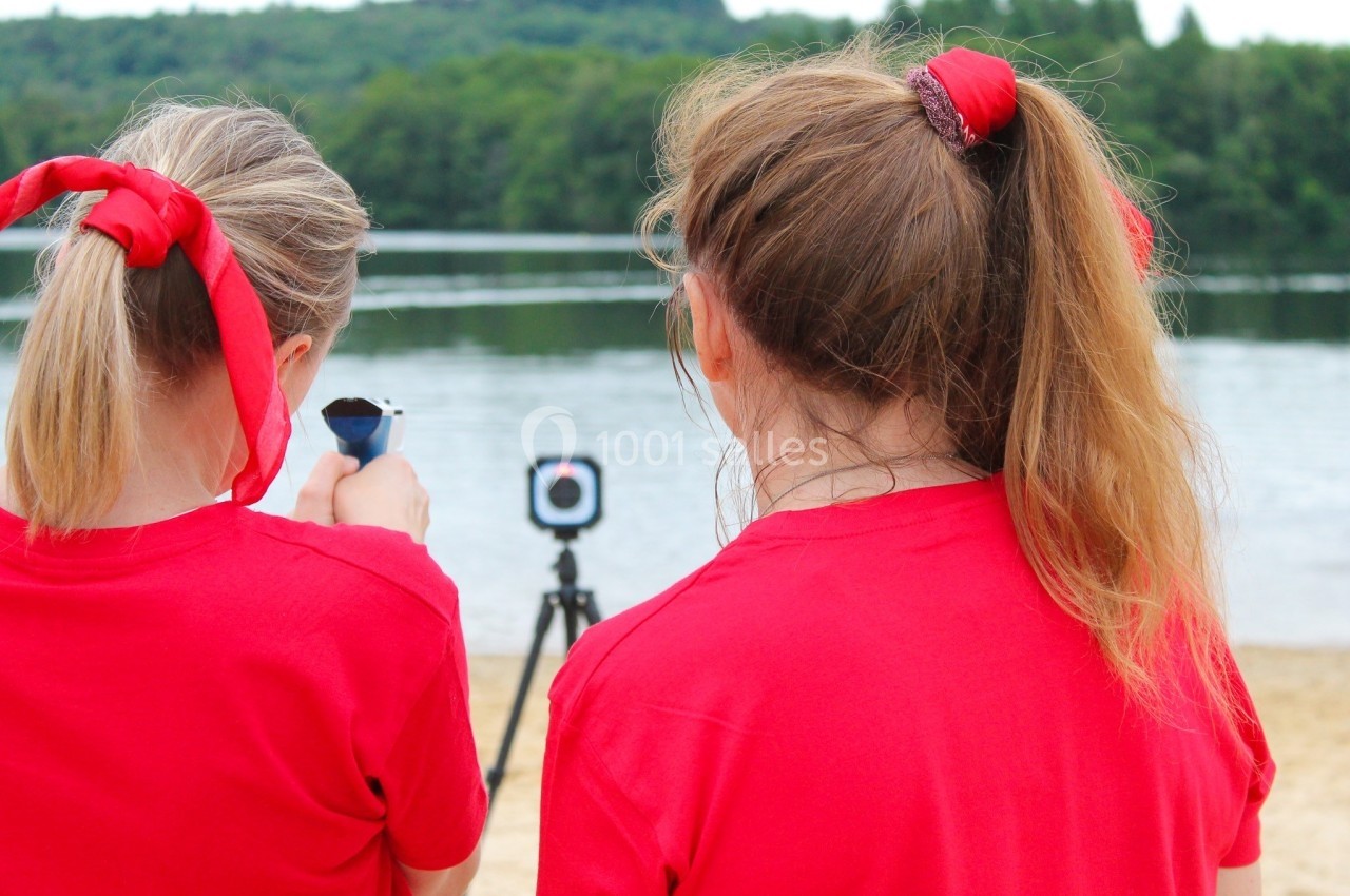 Deux personnes en t-shirts rouges observent un lac, l'une tenant un appareil, avec un trépied et une caméra en arrière-plan.