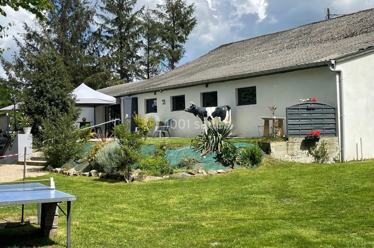 Jardin avec pelouse, table de ping-pong, plantes, statue de vache et maison blanche en arrière-plan.
