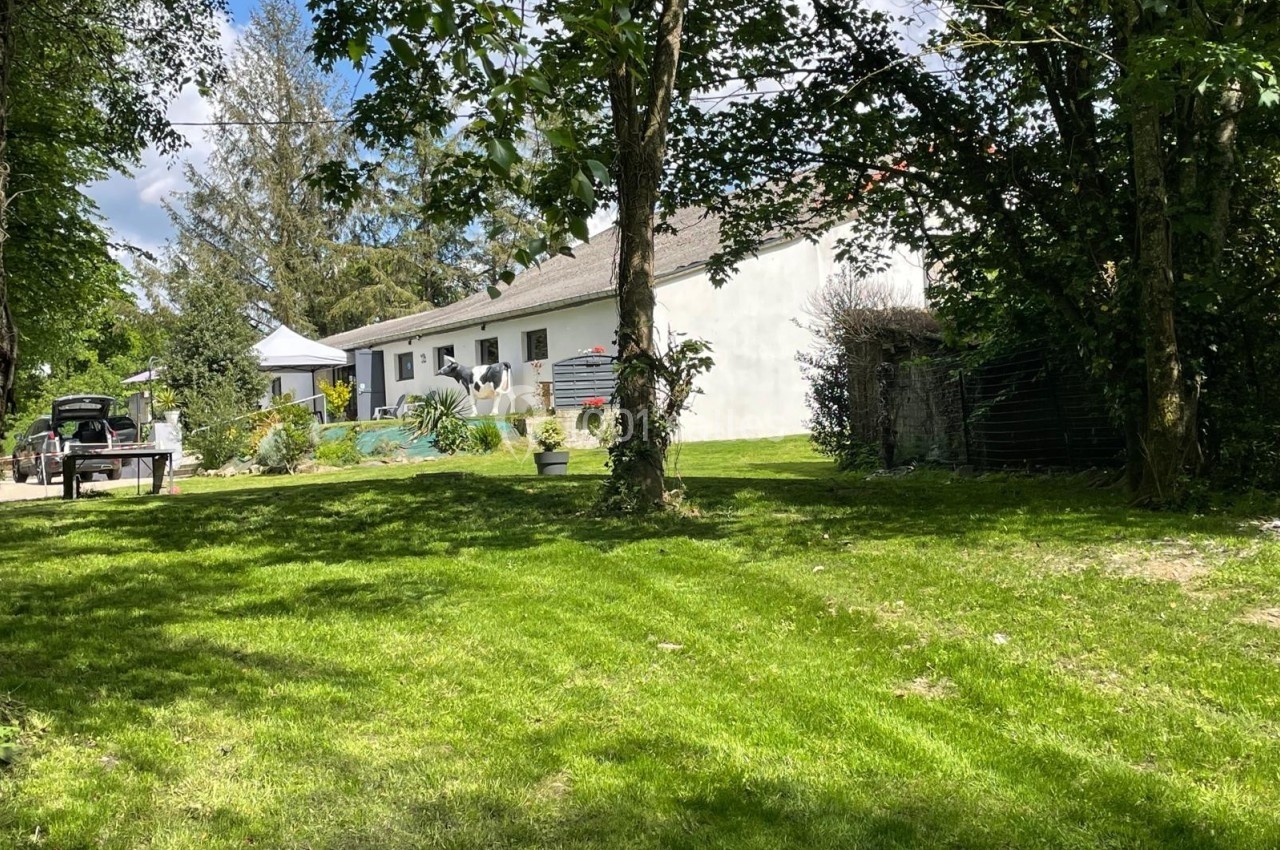 Maison blanche entourée de verdure, avec pelouse, arbres et terrasse visible sous un ciel partiellement dégagé.