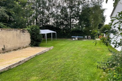 Maison moderne éclairée la nuit, entourée d'un jardin avec des arbustes et des fleurs.