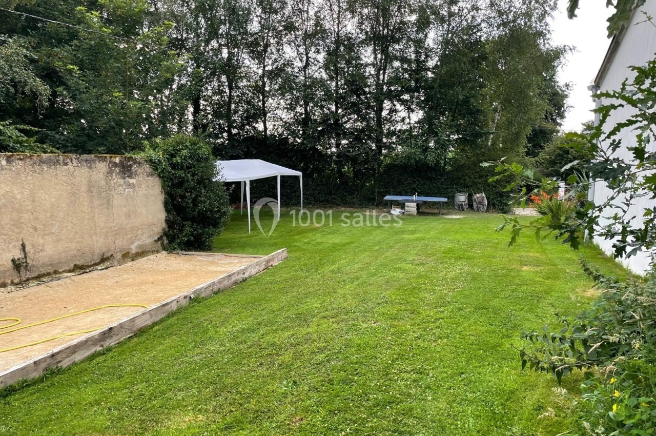 Jardin avec pelouse, tente blanche, table et chaises près d'un mur en pierre et d'une haie d'arbres.