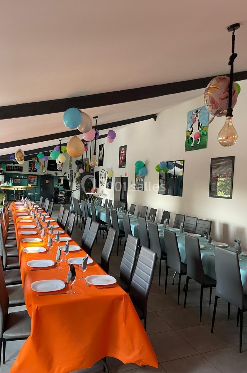 Salle décorée avec des ballons colorés, tables alignées avec nappes orange et turquoise, préparées pour un repas.