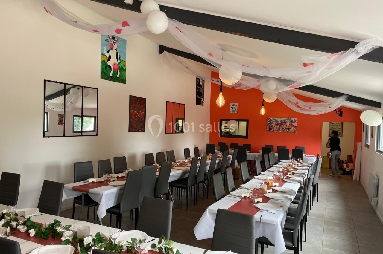 Salle décorée pour un repas festif, avec longues tables dressées, nappes rouges et blanches, et guirlandes suspendues.