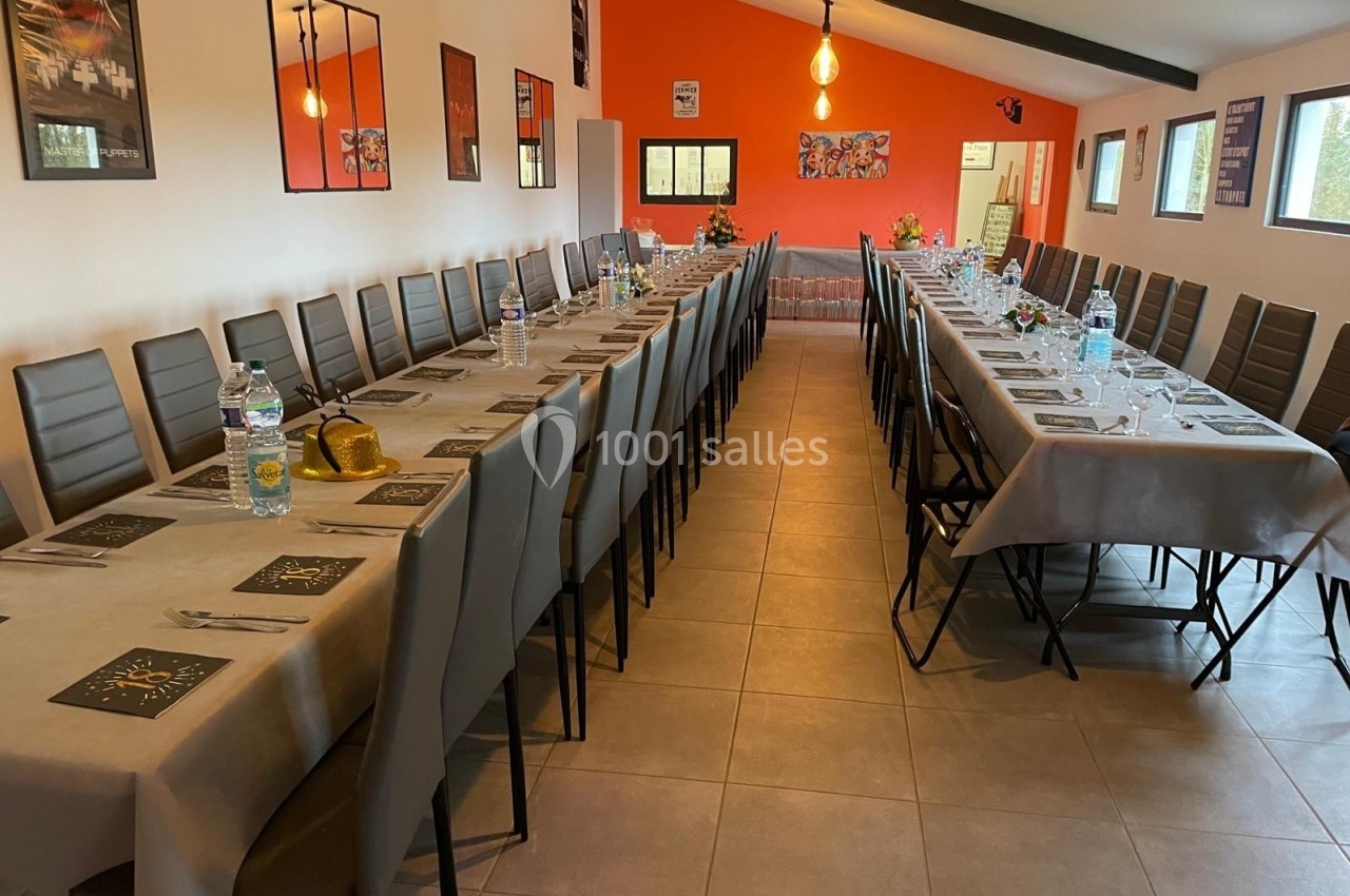 Salle aménagée avec deux longues tables dressées, chaises alignées, murs orange et blanc, éclairage suspendu.