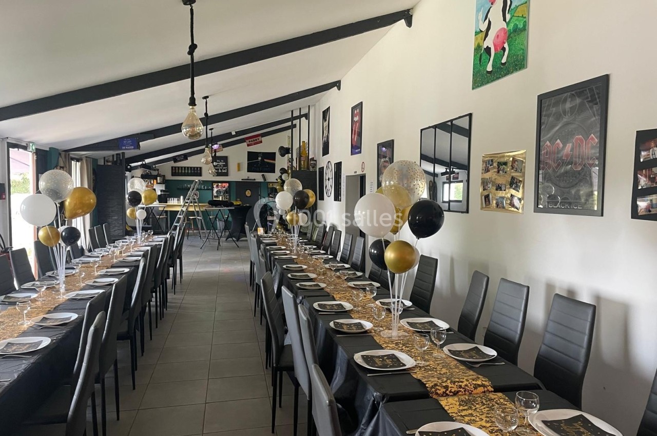 Salle décorée pour un événement avec longues tables dressées, nappes noires, ballons dorés et blancs, et décorations murales.