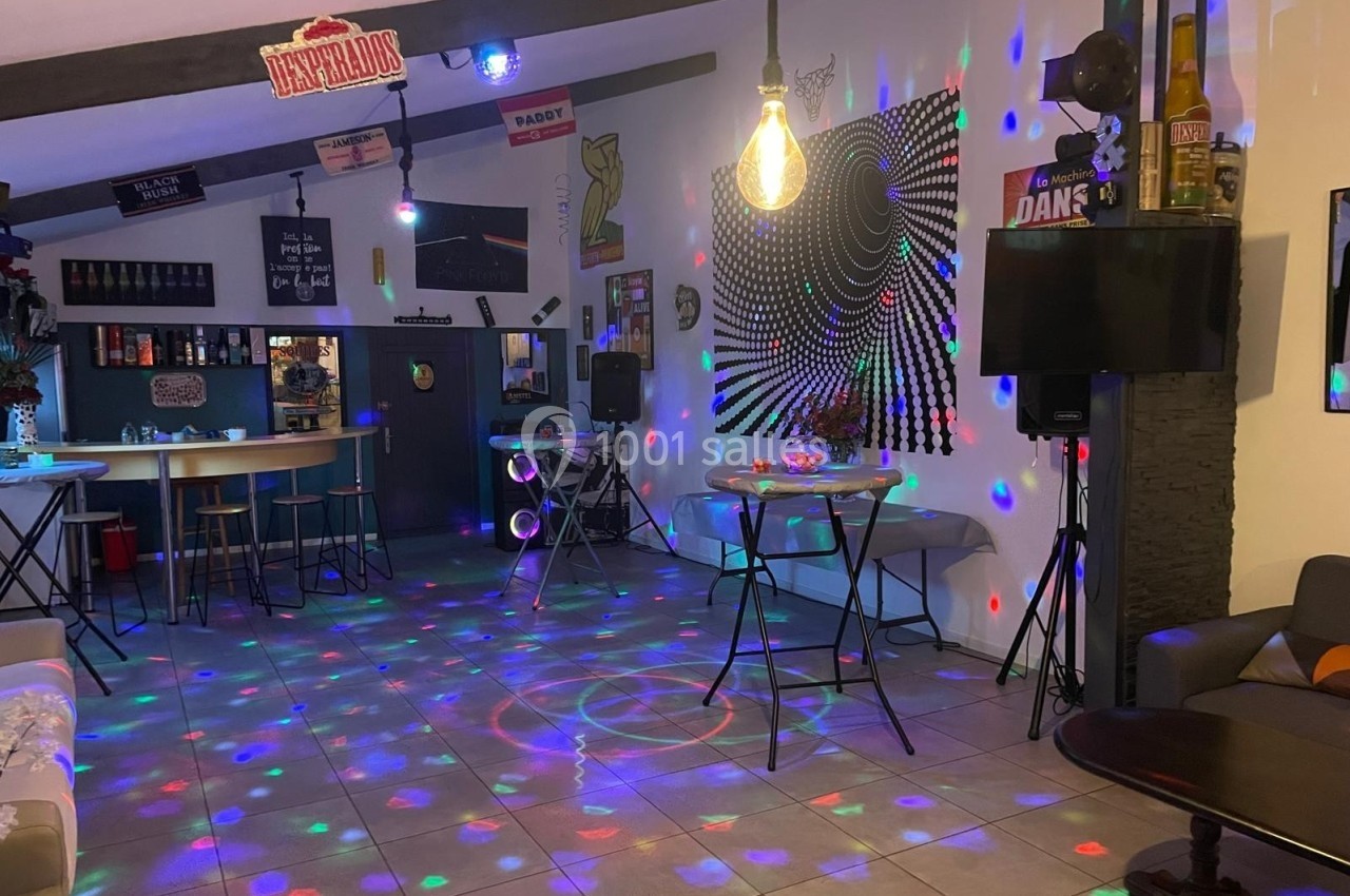 Salle décorée avec des jeux de lumière colorés, tables hautes et ambiance festive, bar en arrière-plan.