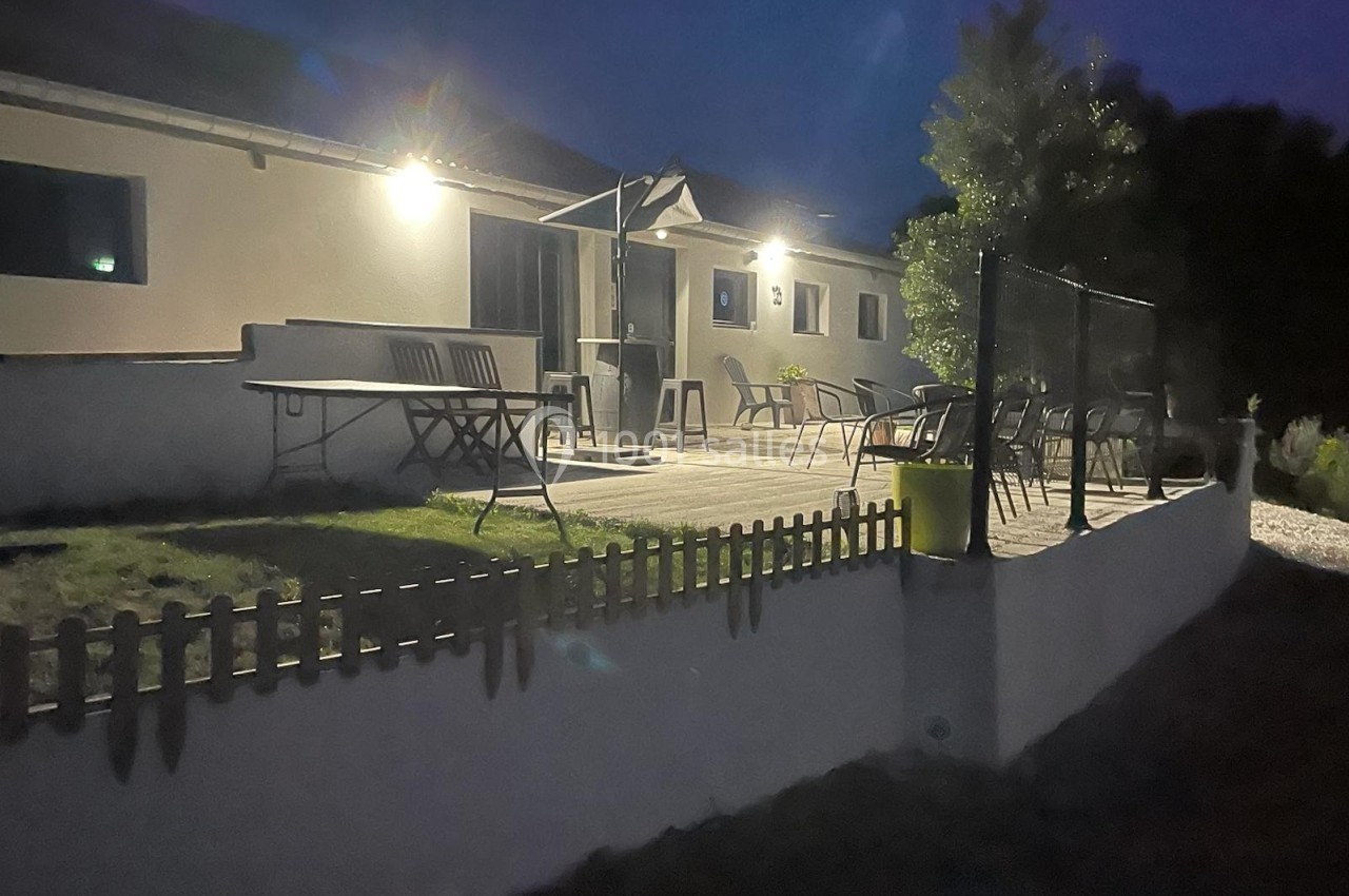 Terrasse éclairée d'une maison la nuit, avec mobilier de jardin, pelouse et petite clôture en bois au premier plan.