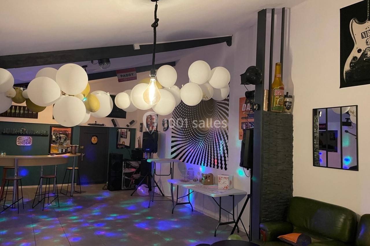 Salle décorée avec des ballons blancs, éclairage tamisé et mobilier de bar, ambiance festive avec jeux de lumière.