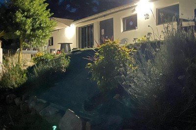 Maison moderne éclairée la nuit, entourée d'un jardin avec des arbustes et des fleurs.