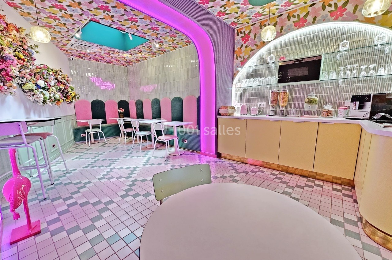 Salle de café colorée avec décor floral, éclairage néon rose et mobilier moderne sur un sol en carreaux.
