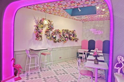 Salle de café colorée avec décoration florale murale, néons roses, mobilier pastel et ambiance lumineuse.