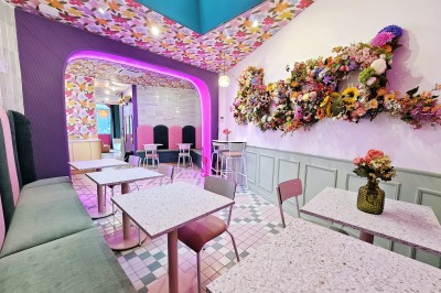 Salle de café colorée avec décoration florale murale, néons roses, mobilier pastel et ambiance lumineuse.
