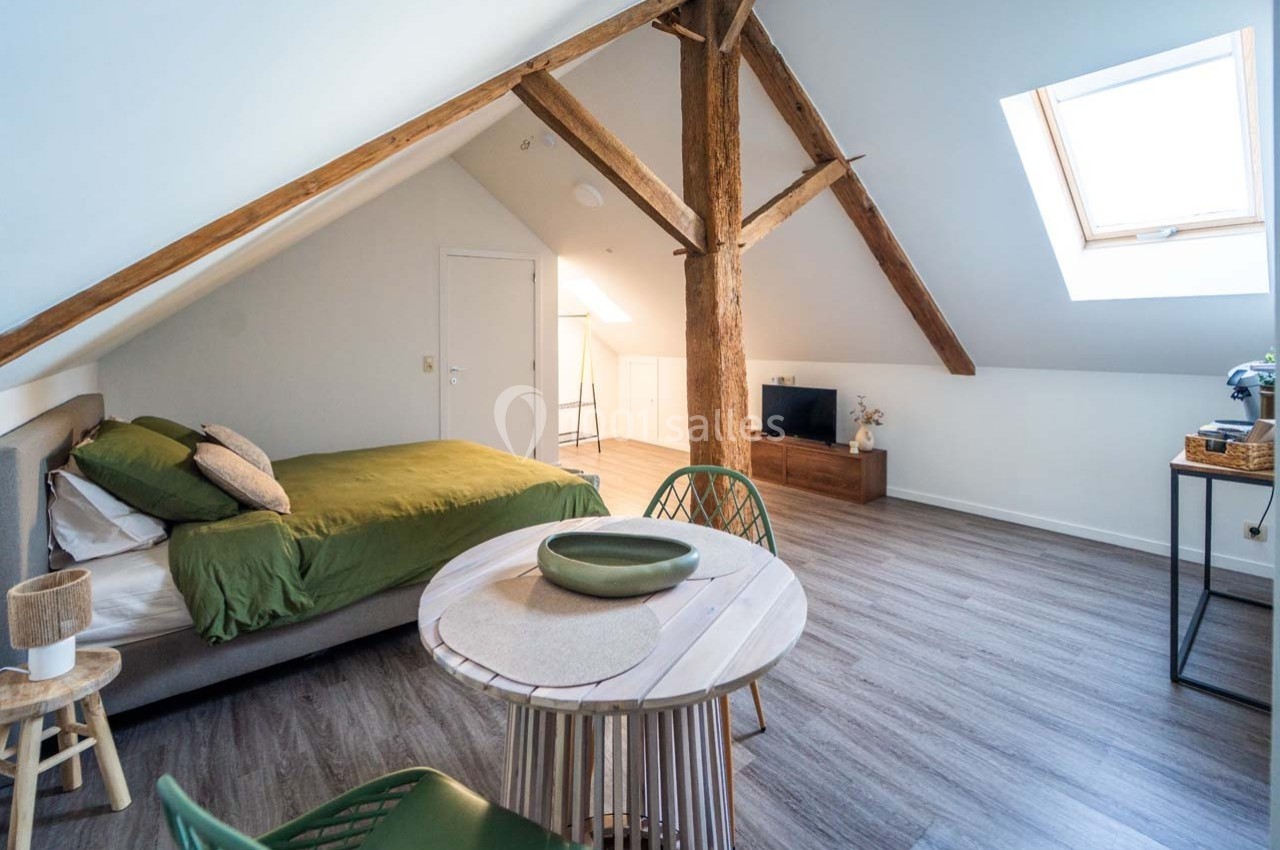 Chambre mansardée lumineuse avec lit double, table ronde, poutres apparentes et fenêtre de toit.